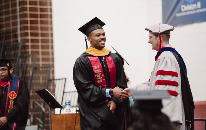 Graduate in cap and gown shaking hands at commencement ceremony after earning degree.