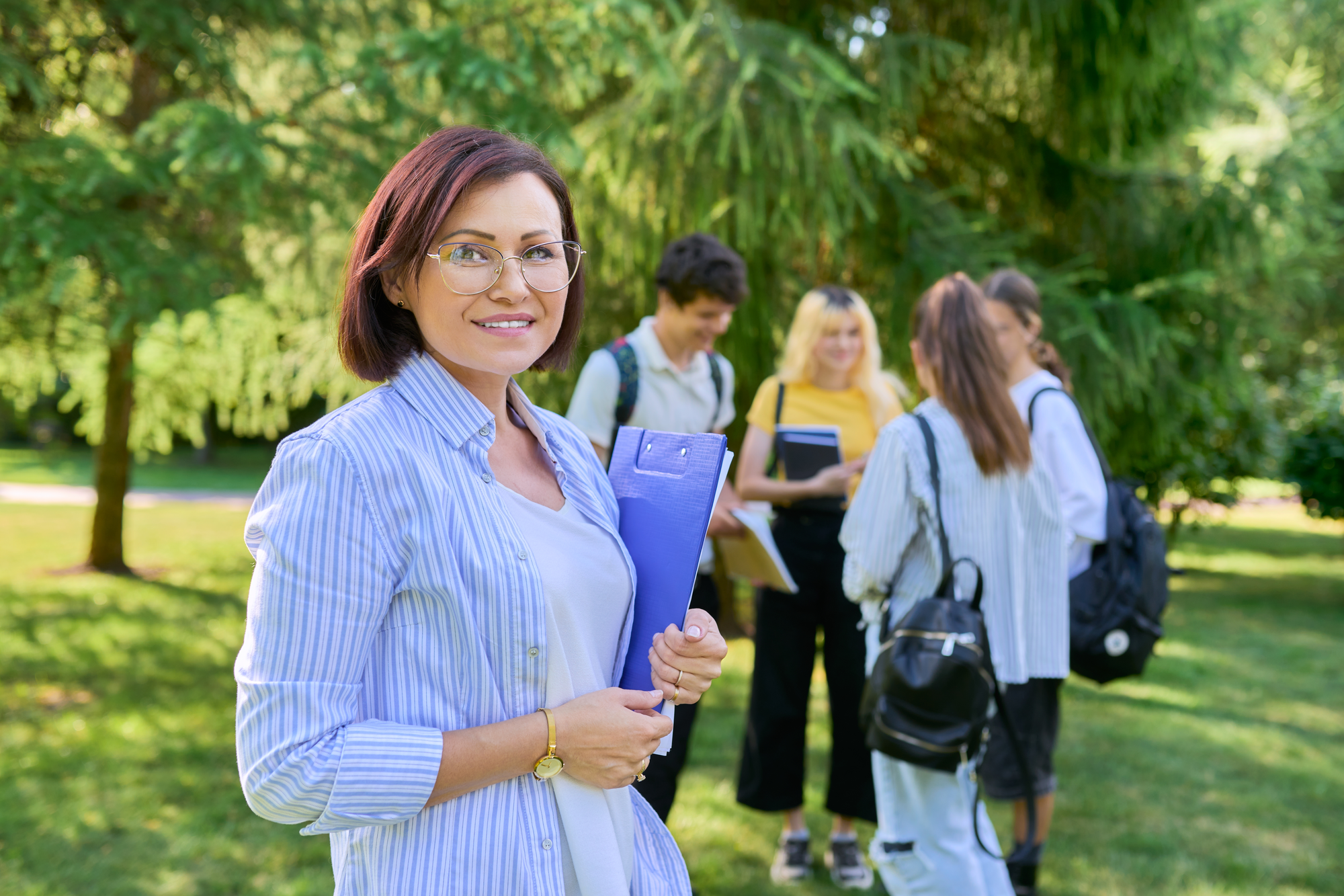 Confident woman holding a folder outdoors with students interacting in the background, symbolizing personal and professional growth through education.